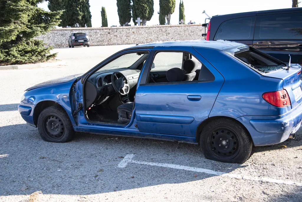 A blue hatchback abandoned in a gravel car park with its doors open and a flat rear tyre