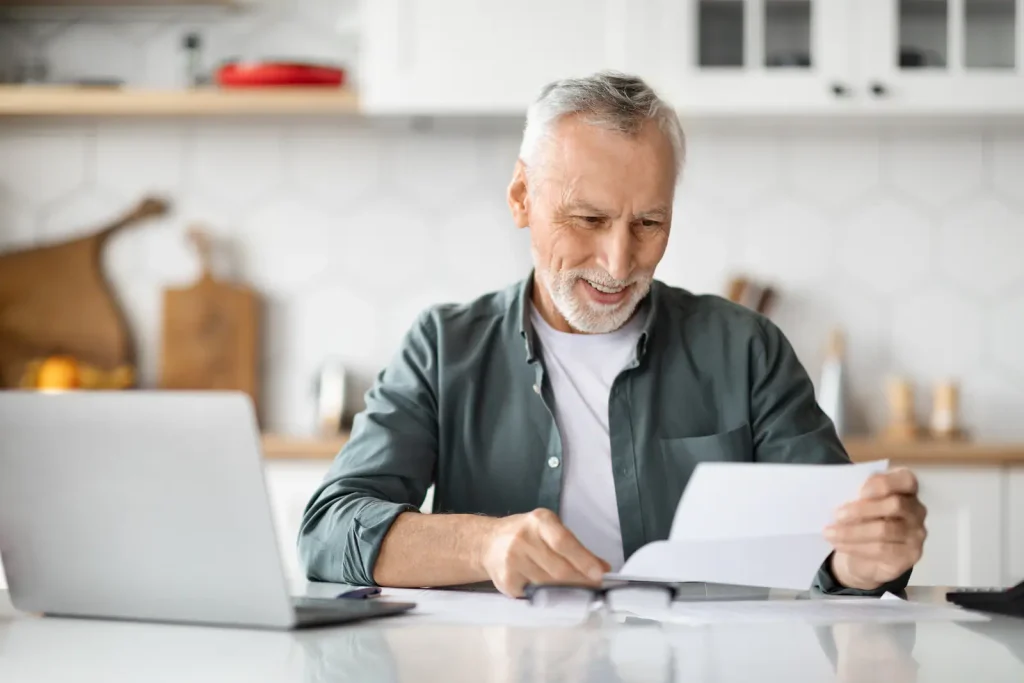 A smiling older man at a kitchen table reading a letter beside an open laptop