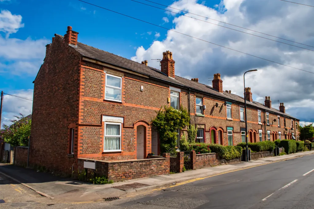 A row of red-brick terraced houses along a quiet road in Manchester under a blue sky